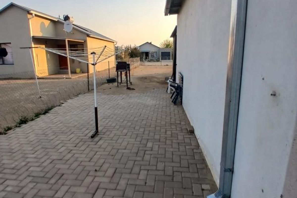 A rear exterior view showing a paved patio area alongside the house wall with a metal downpipe and surrounding fencing.