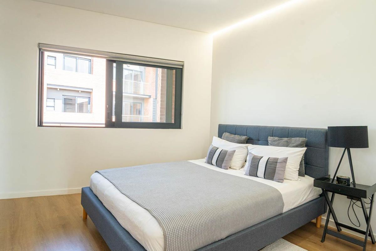 A modern, well-lit bedroom featuring laminate flooring, neutral walls, and a large sliding window providing natural light.