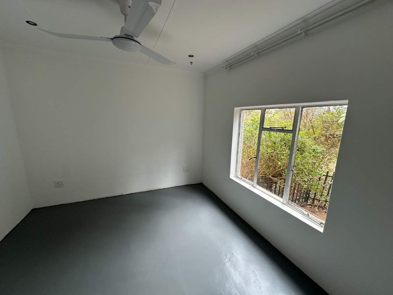 A clean, empty bedroom featuring a ceiling fan, painted concrete flooring, and a large window with views of the surrounding greenery.
