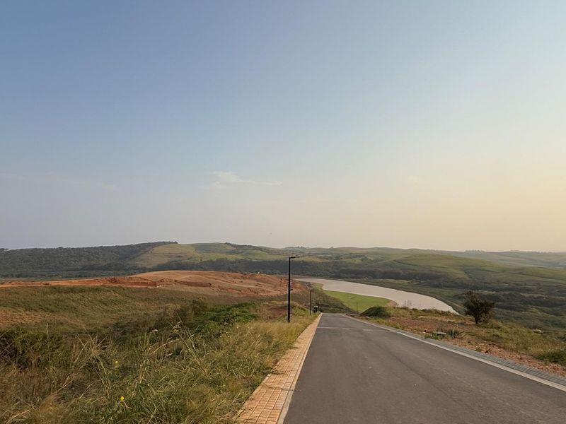 A view of a paved residential road leading through a developing landscape with rolling hills and a nearby river.