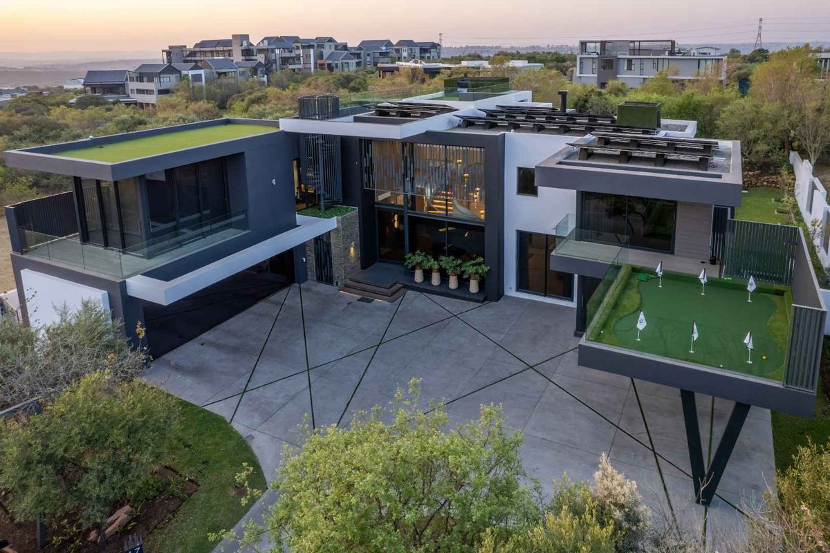Aerial view of a modern luxury residence featuring a concrete driveway, integrated putting green, and contemporary architectural design with flat roof sections.