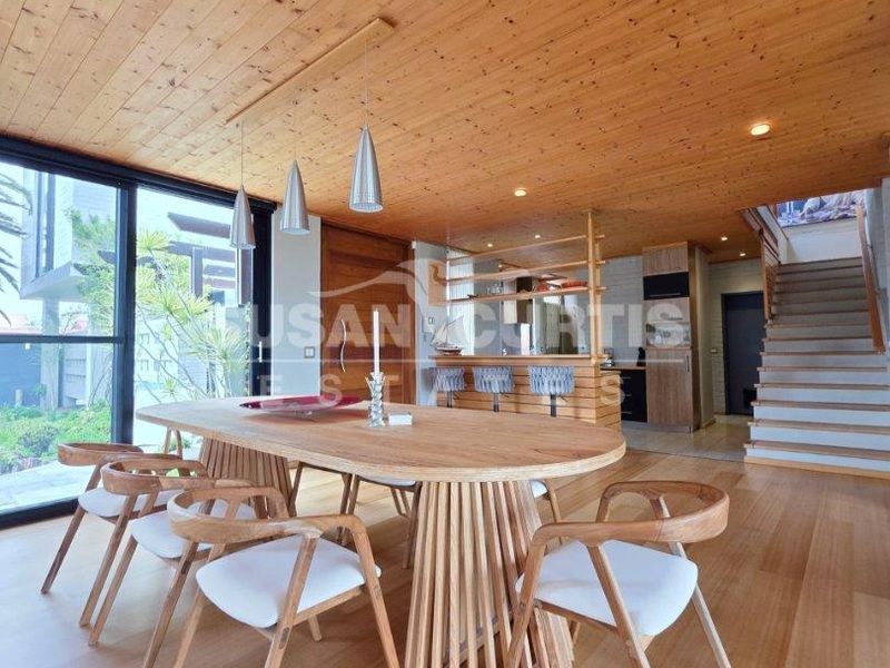 Spacious open-plan living and dining area featuring warm timber ceilings and flooring, large glass doors, and a modern integrated kitchen with staircase access.