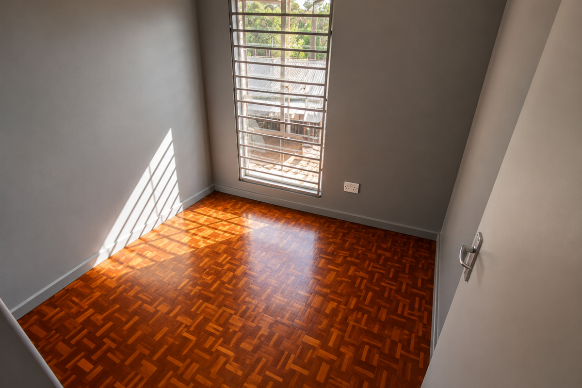 An empty bedroom featuring well-maintained parquet flooring, neutral grey walls, and a window fitted with security bars.