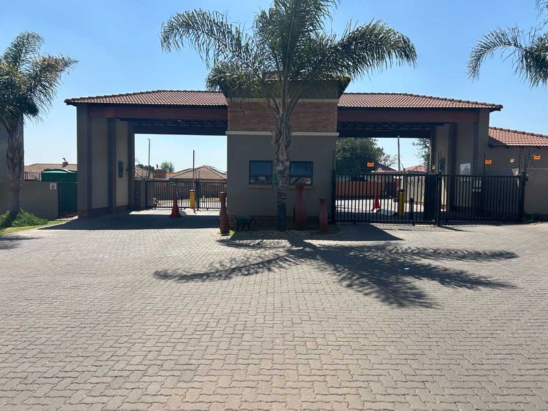 The main entrance to the townhouse complex featuring a secure gated access point, guard house, and paved driveway.