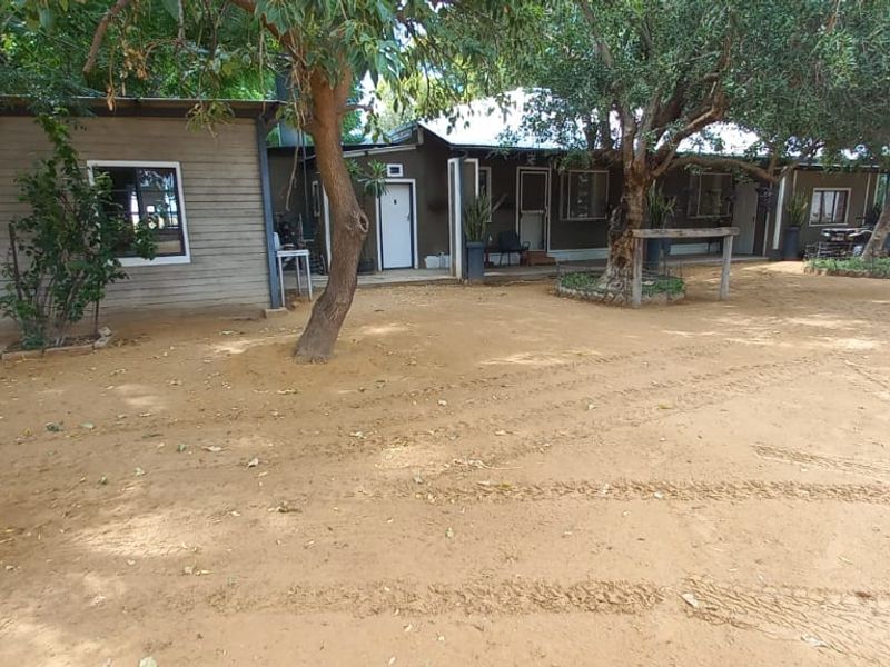 Rear view of the farm dwelling featuring timber siding, a corrugated metal roof, and a covered patio area set within a sandy yard.