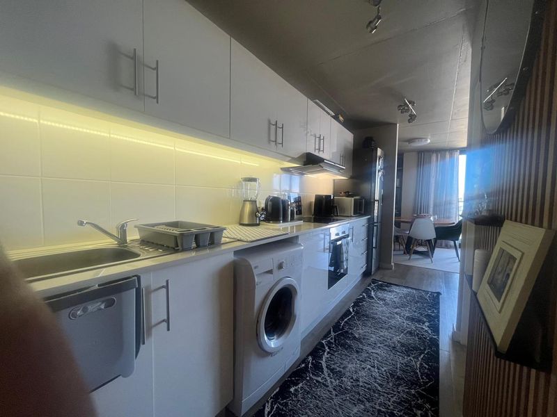 A modern galley-style kitchen featuring white cabinetry, integrated appliances, under-cabinet lighting, and a tiled backsplash, leading into an open-plan living and dining area.
