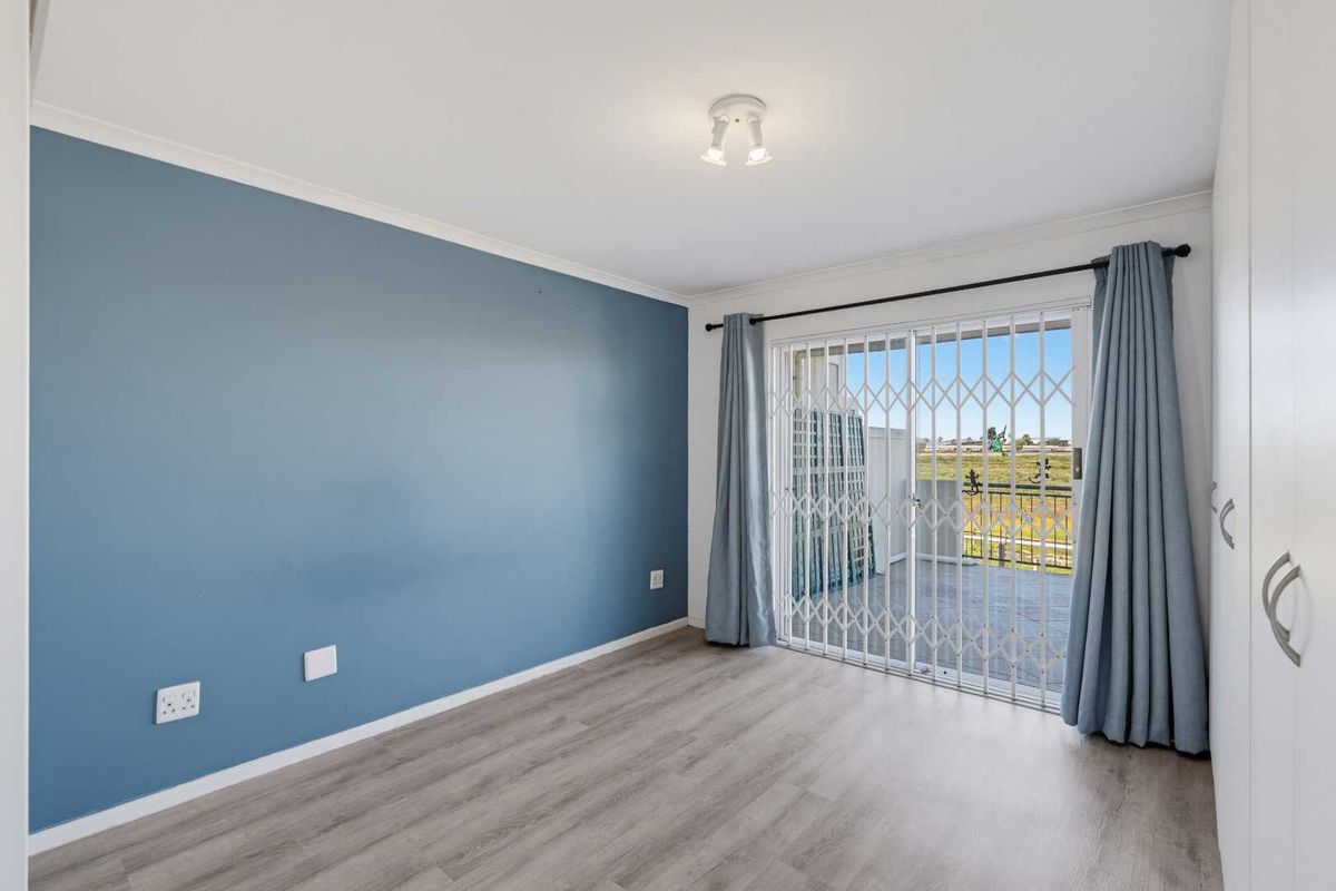 A bright, empty bedroom featuring modern laminate flooring, a blue accent wall, built-in cupboards, and a sliding door leading to a private balcony.