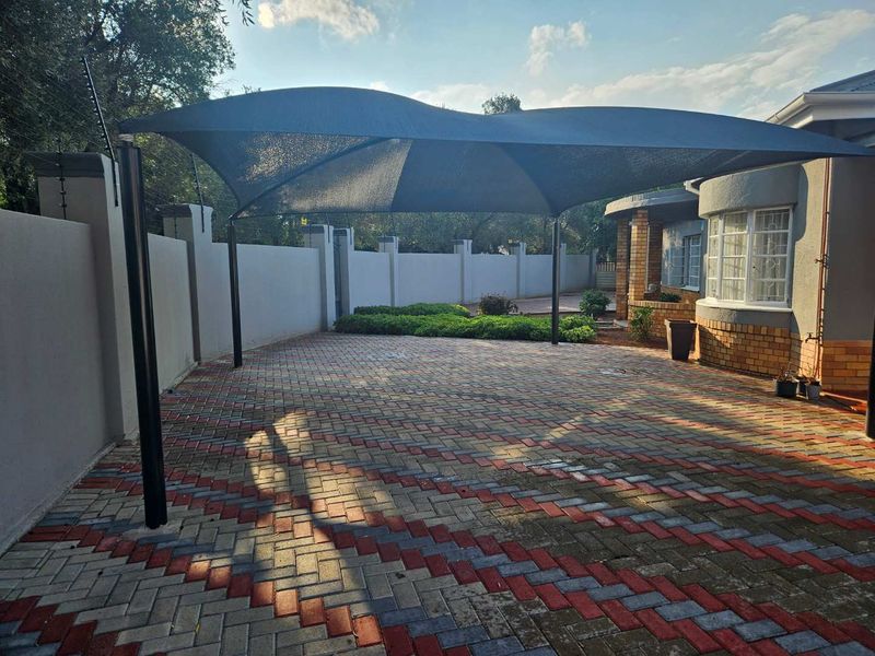 A paved front driveway area featuring a large shade sail, perimeter wall with electric fencing, and a brick-accented building facade.