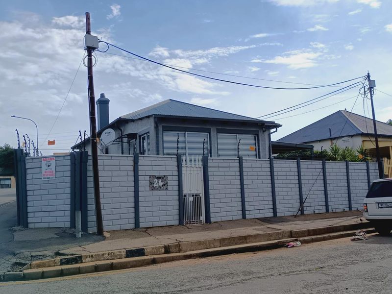 Front exterior view of a residential property featuring a secure perimeter wall with electric fencing, metal gate, and a corrugated metal roof.