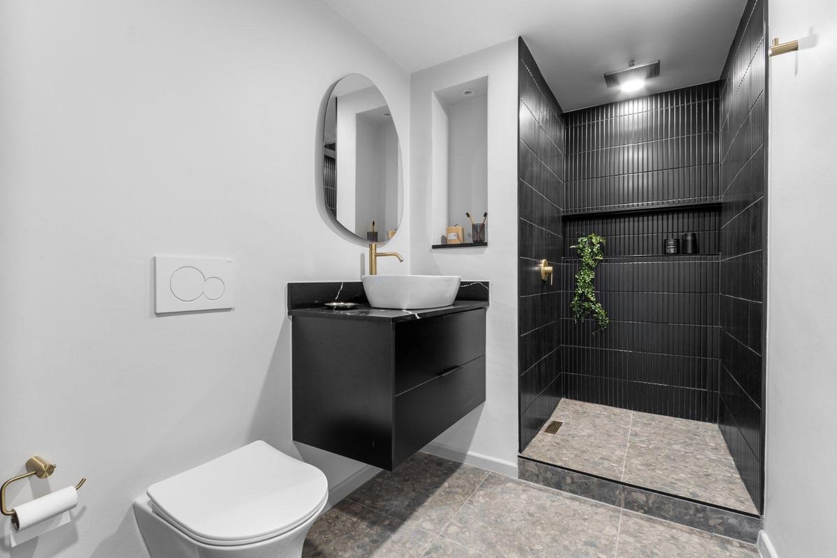 Modern bathroom featuring a floating vanity with vessel sink, wall-mounted toilet, and a sleek walk-in shower with dark vertical tiling and brass accents.