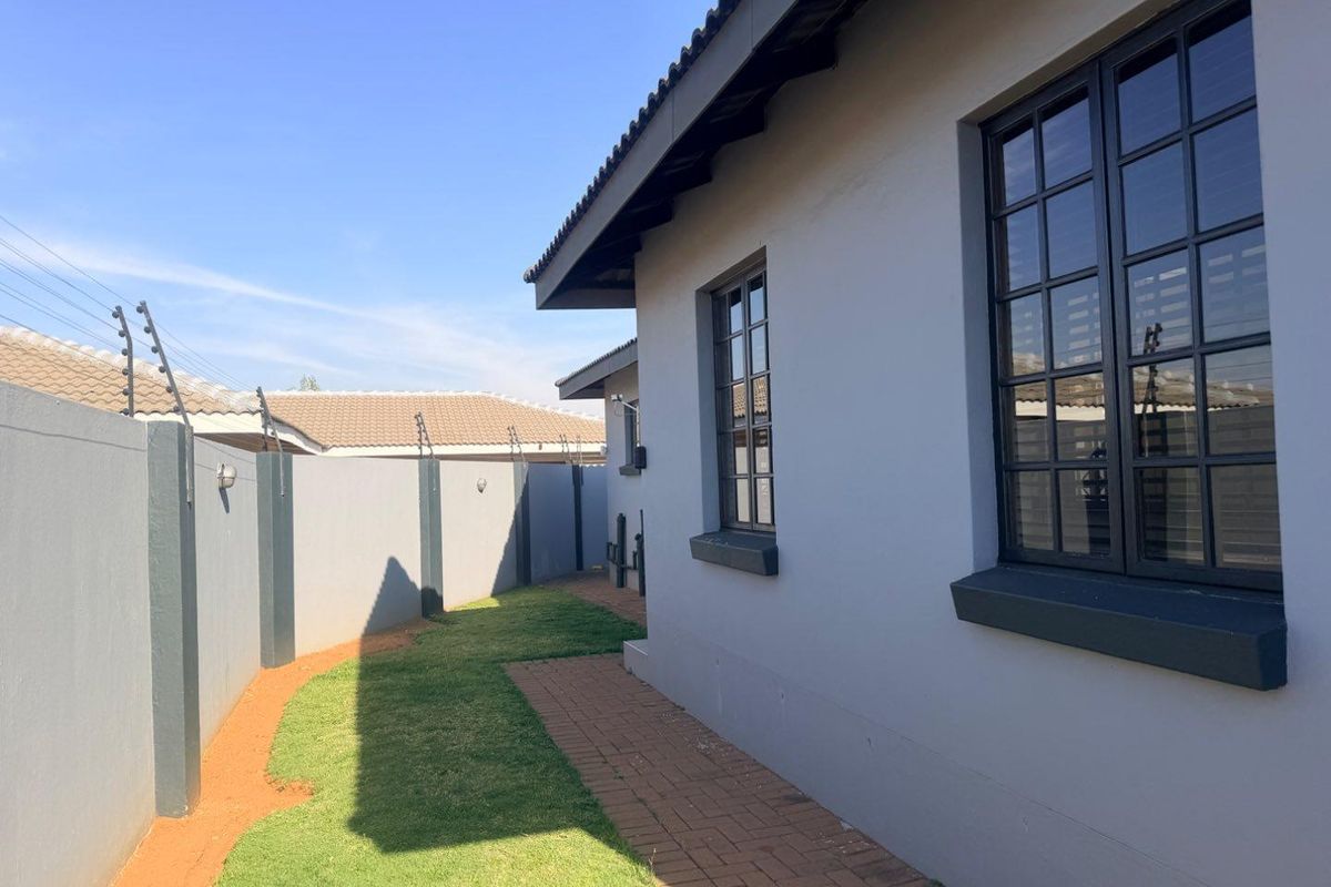 A well-maintained rear exterior view featuring a paved walkway, manicured lawn, and secure perimeter wall with electric fencing.