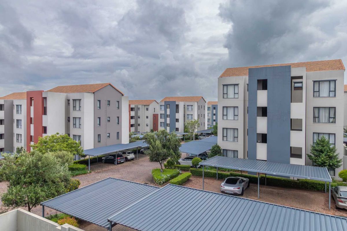 An elevated view of a modern apartment complex featuring multi-story residential buildings, paved parking areas, and covered carports within a landscaped estate.