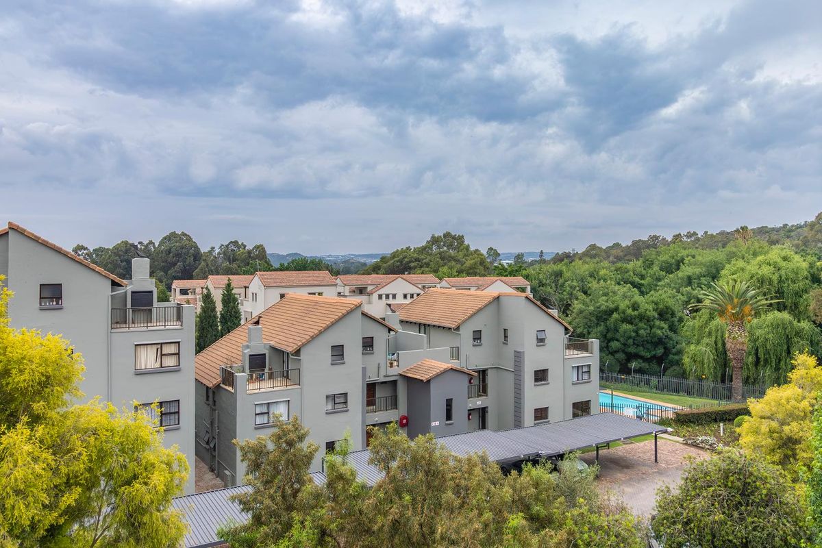 An elevated aerial view of the apartment complex, featuring multi-story residential buildings, tiled roofs, covered parking bays, and a communal swimming pool area.