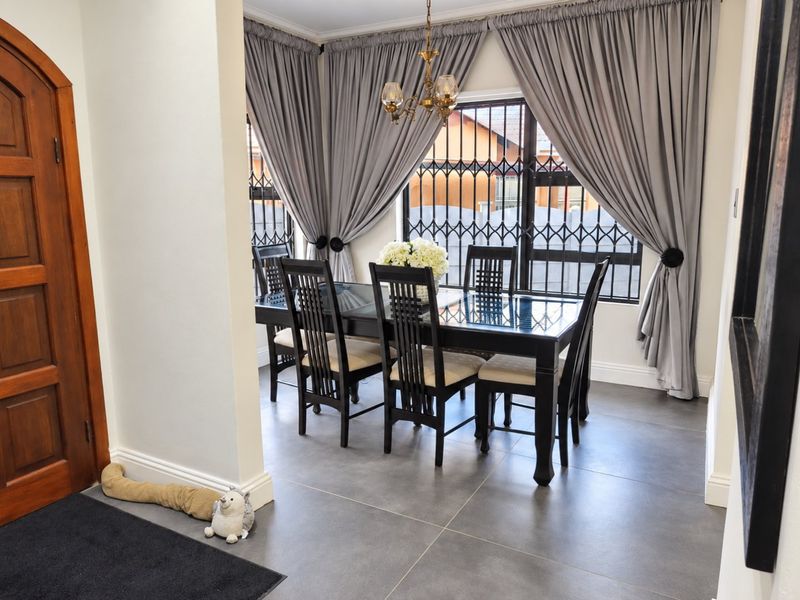 A well-maintained dining area featuring large format floor tiles, decorative chandelier lighting, and windows fitted with security bars.