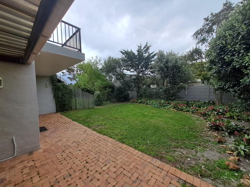A rear garden view featuring a brick-paved patio area, a lawn, established garden beds, and an elevated balcony with metal railings.