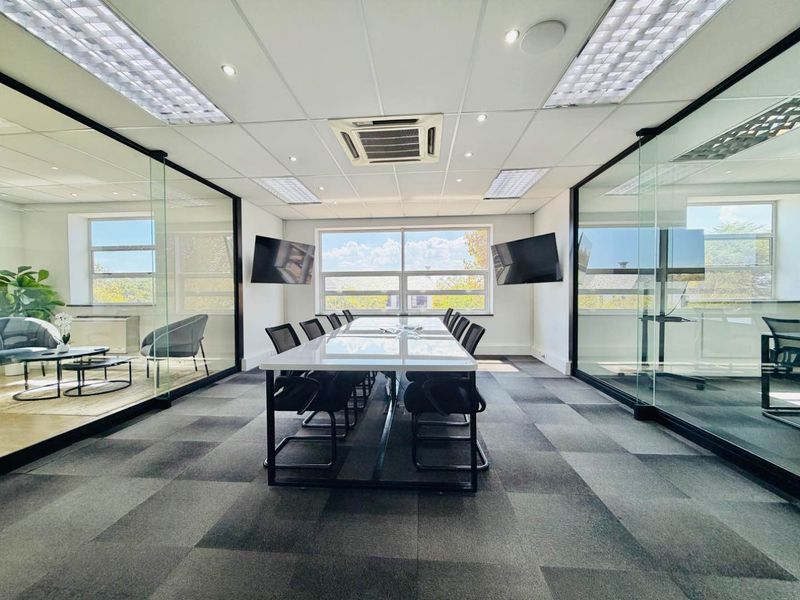Modern professional boardroom featuring glass partition walls, carpeted flooring, recessed lighting, and integrated air conditioning in a bright, contemporary office environment.