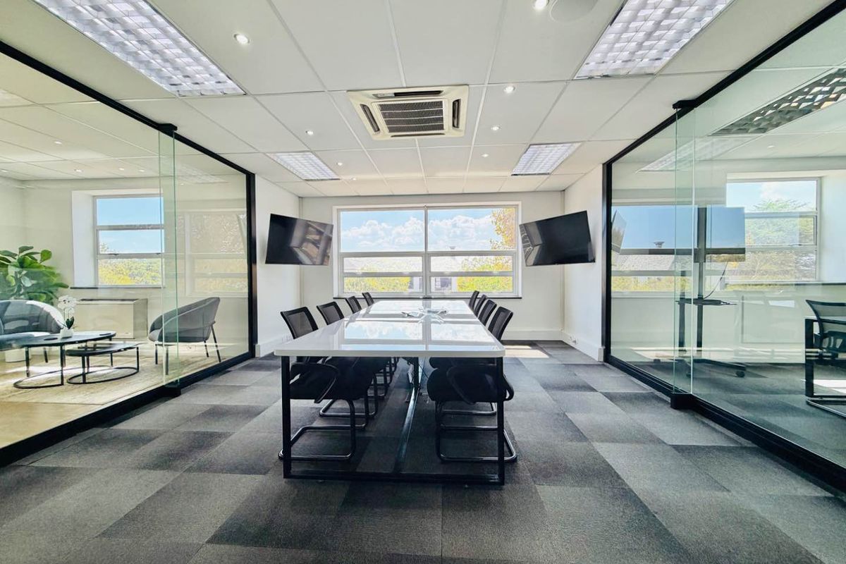 Modern professional boardroom featuring glass partition walls, carpeted flooring, recessed lighting, and integrated air conditioning in a bright, contemporary office environment.