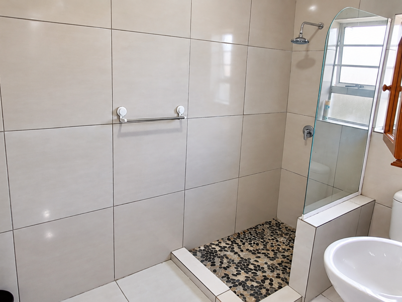 A clean bathroom featuring a walk-in shower with a pebble floor, glass screen, pedestal basin, and toilet, finished with neutral wall tiling.