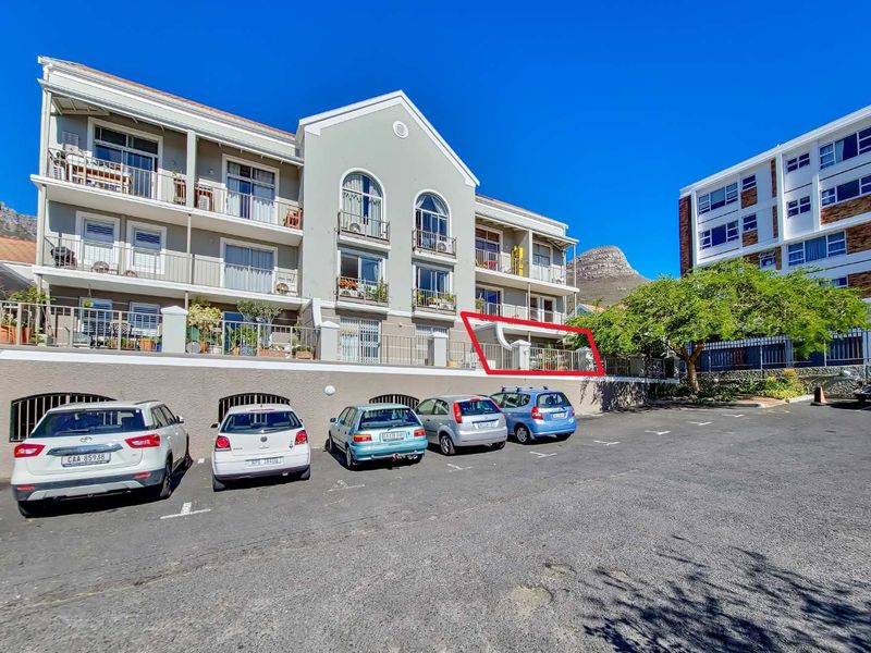 Exterior view of an apartment building featuring a highlighted ground-floor balcony with metal railings and sliding glass doors, situated above a paved parking area.
