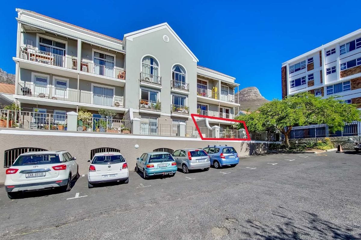 Exterior view of an apartment building featuring a highlighted ground-floor balcony with metal railings and sliding glass doors, situated above a paved parking area.