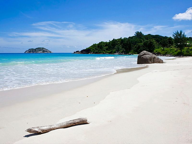 A scenic view of a pristine white sand beach with turquoise ocean waters, lush coastal vegetation, and large granite boulders.