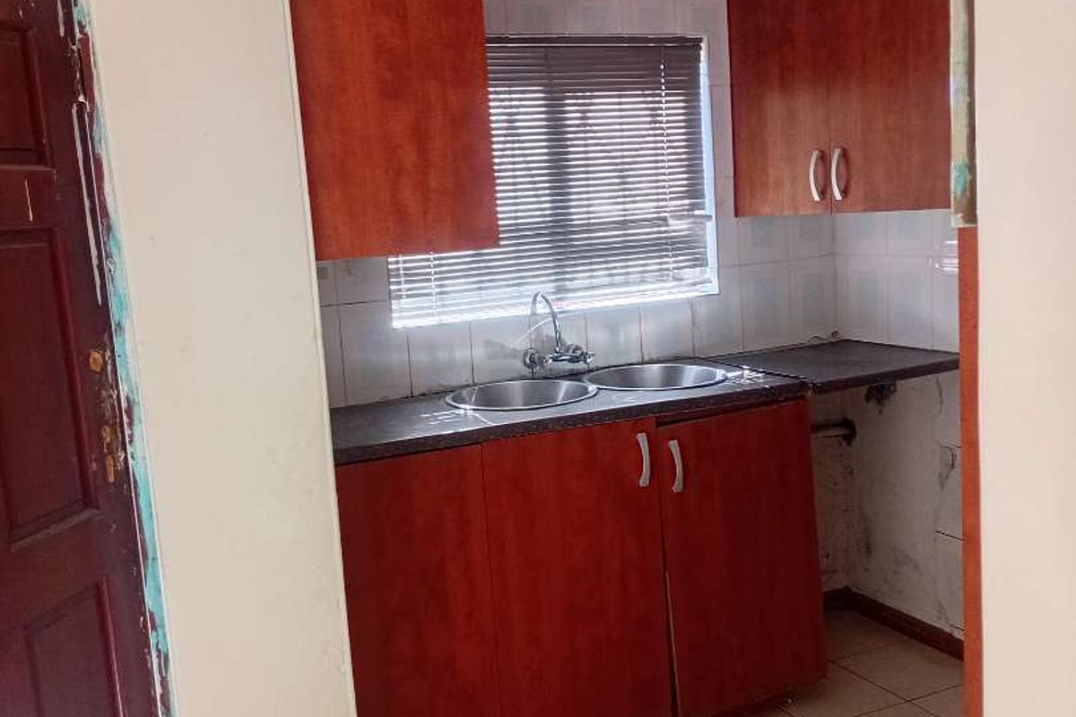 A compact kitchen featuring wooden cabinetry, a double bowl sink, and tiled flooring, currently in need of maintenance and cosmetic updates.