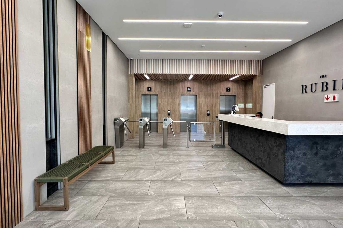 Modern office building lobby featuring a reception desk, secure turnstile access control, and a bank of elevators with contemporary timber and stone finishes.