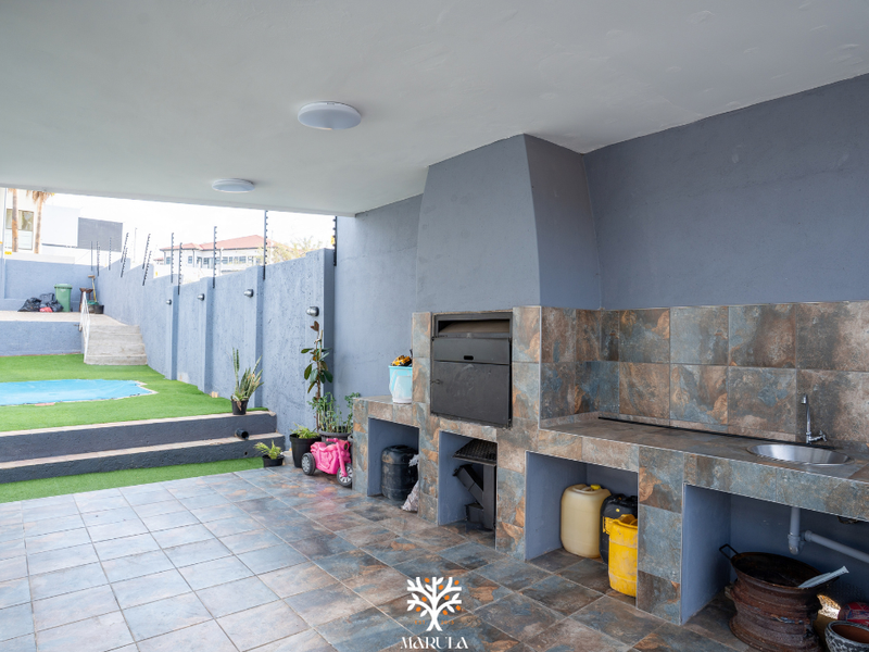 Covered patio area featuring a built-in braai, tiled workspace with integrated sink, and direct access to a tiered garden with artificial lawn.
