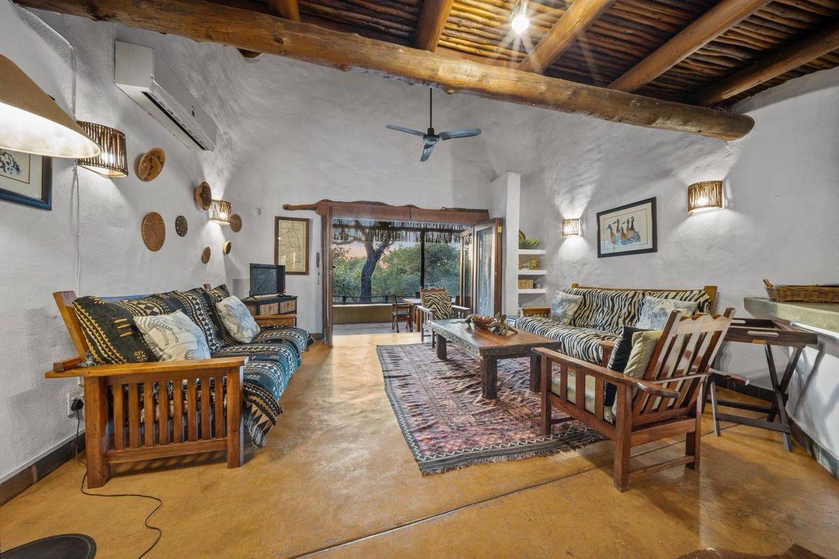 A rustic living room featuring exposed timber ceiling beams, textured white walls, polished concrete flooring, and a ceiling fan, opening onto a patio area.