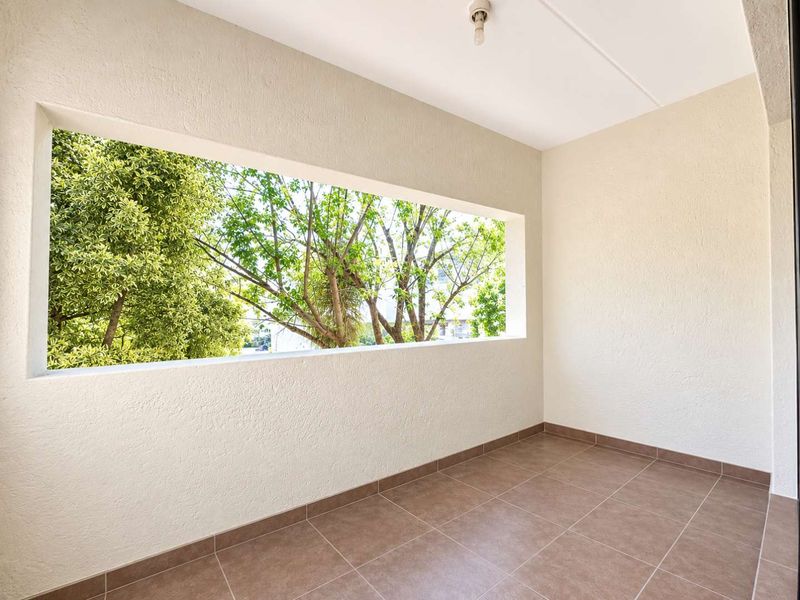 A clean, empty balcony featuring tiled flooring, textured walls, and a large open window frame overlooking mature trees.