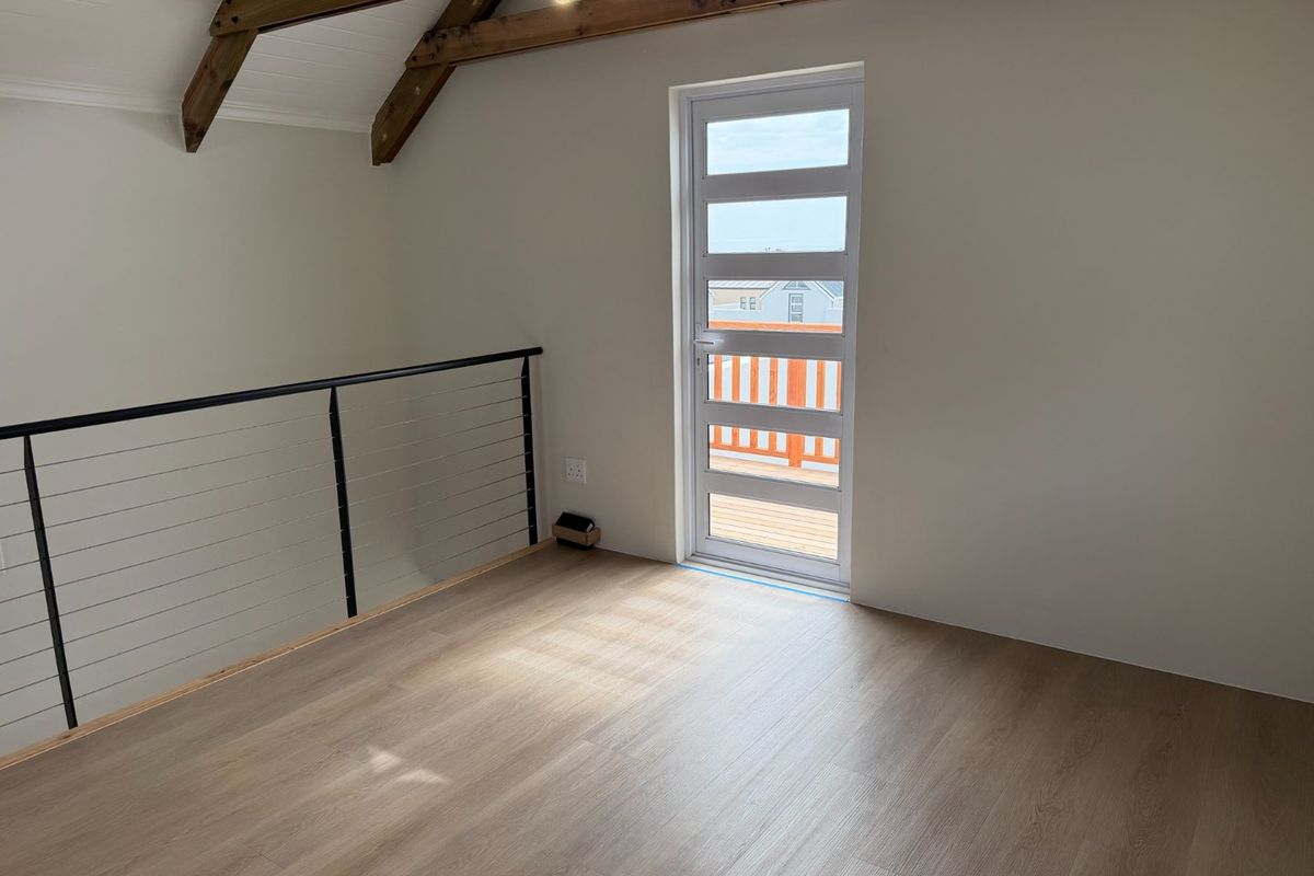 A bright, empty bedroom featuring exposed timber roof trusses, modern laminate flooring, a sleek metal balustrade, and a glass door leading to a balcony.