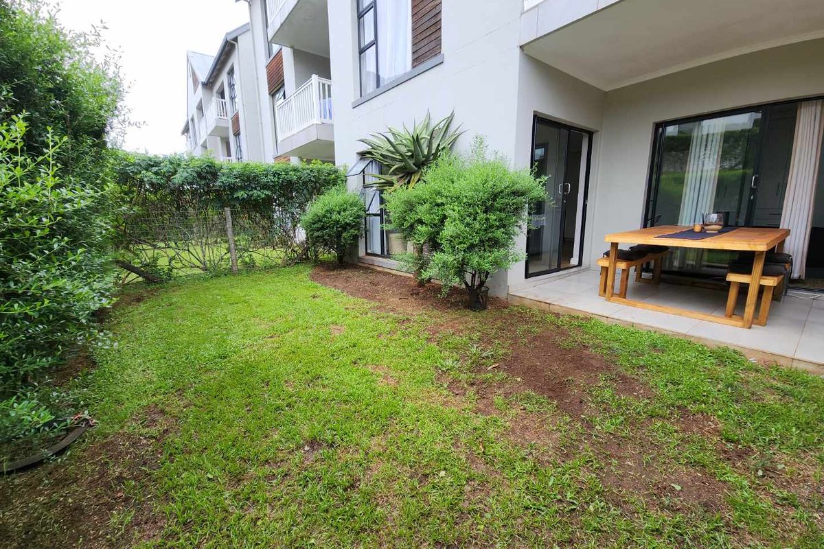 Rear exterior view of a townhouse featuring a private garden with lawn, hedging, and a paved patio area accessible via sliding glass doors.