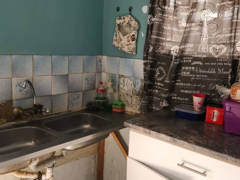 A dated kitchen area featuring a double bowl stainless steel sink, tiled splashback, and laminate countertops, currently showing signs of wear and missing wall tiles.