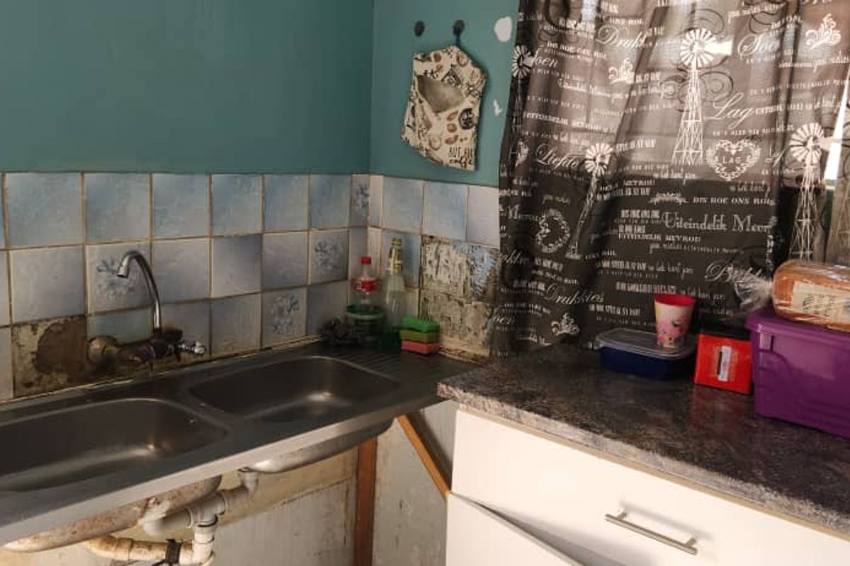 A dated kitchen area featuring a double bowl stainless steel sink, tiled splashback, and laminate countertops, currently showing signs of wear and missing wall tiles.