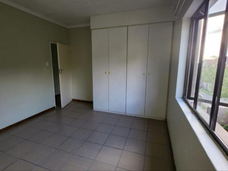 A bright, empty bedroom featuring tiled flooring, built-in cupboards, and a large window providing natural light.