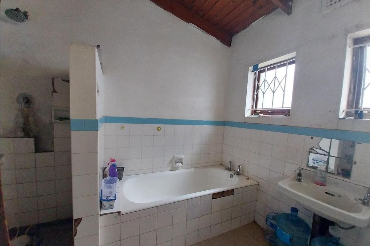 A dated bathroom featuring a built-in bathtub, wall-mounted shower, and pedestal basin, characterized by exposed timber ceiling beams and original tiled finishes.