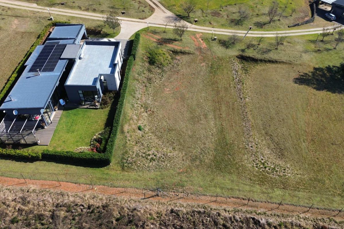 Aerial view of a vacant residential plot adjacent to an existing modern home, featuring open grass land and established boundary fencing.