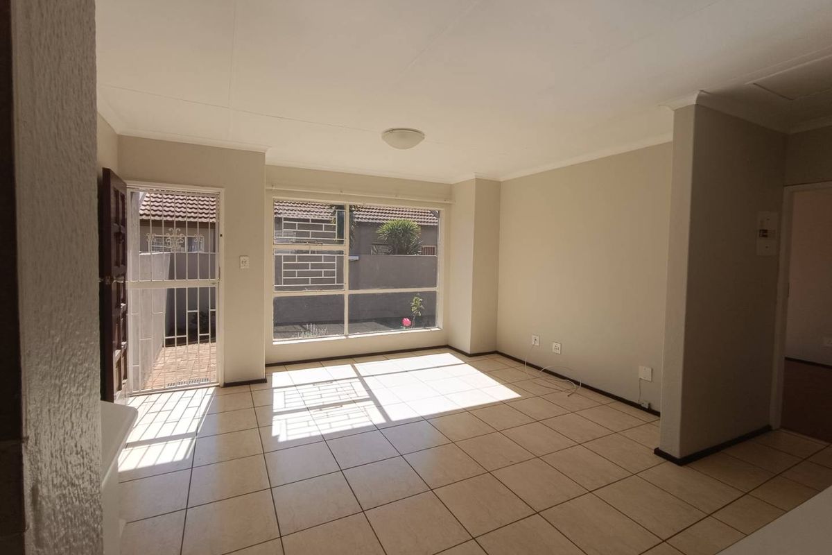 A bright, empty living room featuring neutral tiled flooring, large windows for natural light, and direct access to an outdoor area via a security gate.