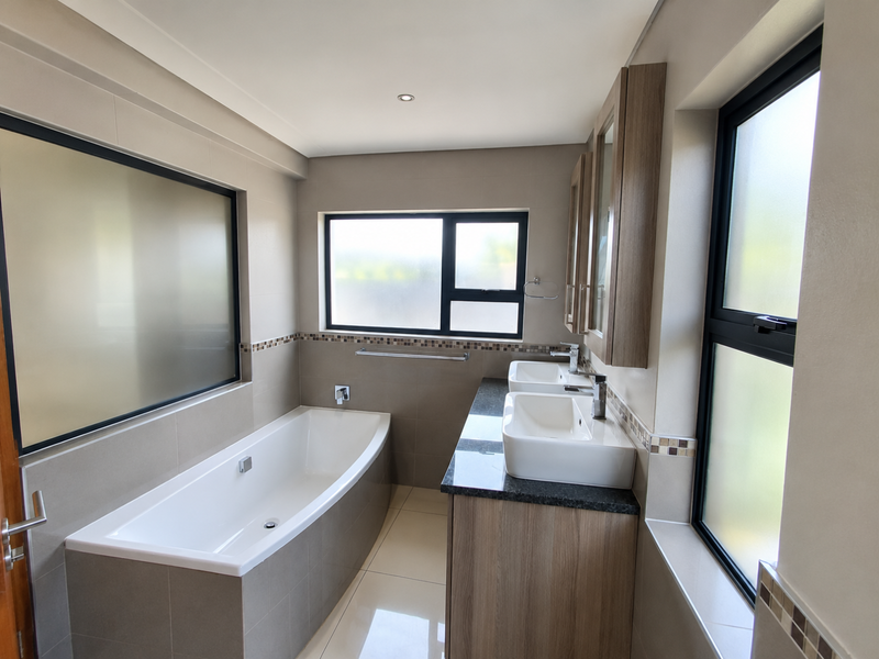 Modern bathroom featuring a built-in bathtub, double vanity with stone countertop, and neutral tiled finishes with frosted glass windows for privacy.