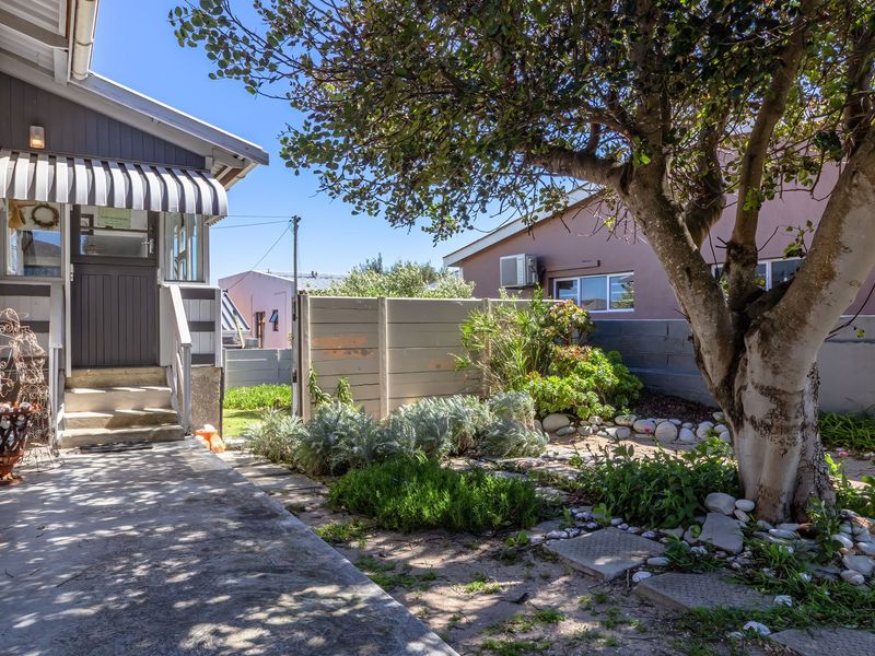 A rear exterior view featuring a concrete pathway, a shaded entrance with a striped awning, and a landscaped garden area with a mature tree.