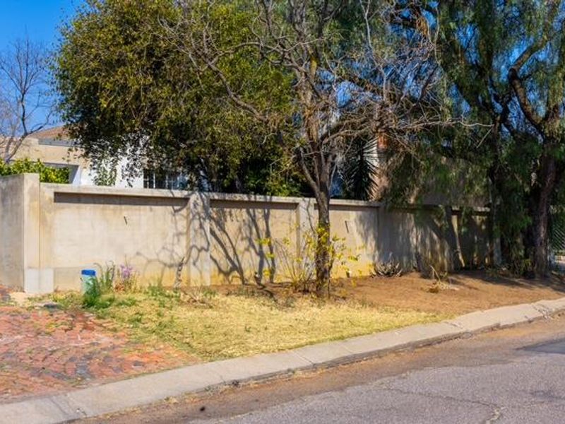 Street view of the property showing a boundary wall, paved driveway access, and mature trees along the roadside.
