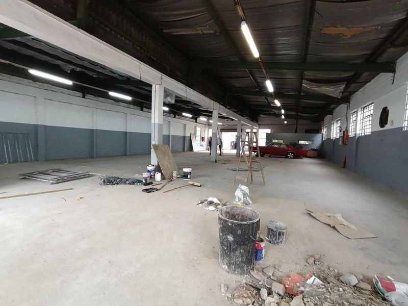 Spacious industrial warehouse area featuring high ceilings with exposed steel trusses, concrete flooring, and ample natural light through industrial-style windows.