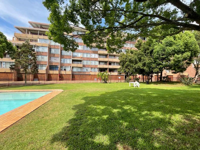 A well-maintained communal pool area featuring a swimming pool, paved surround, and expansive lawn set against the apartment building facade.