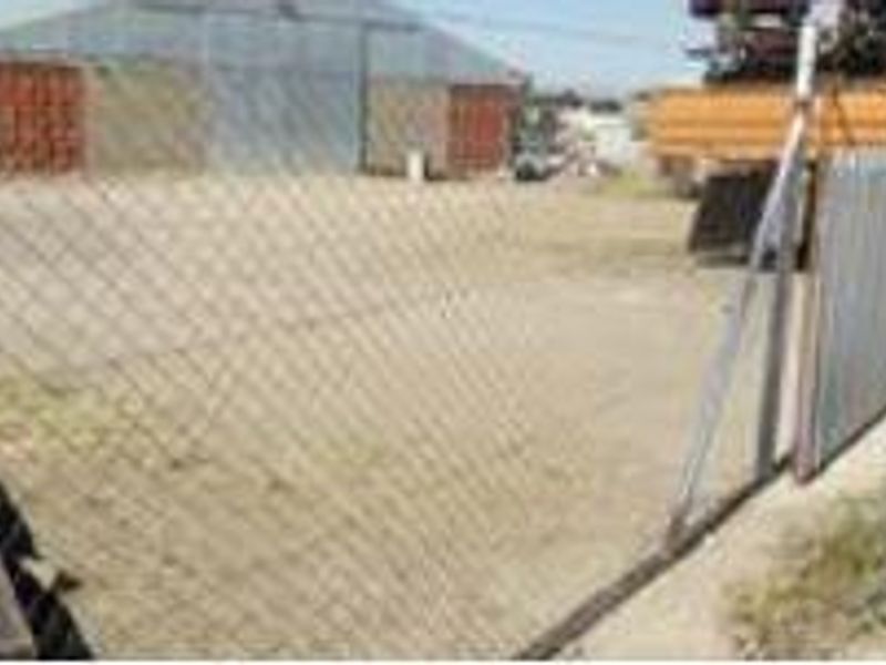 An industrial yard area featuring a large metal shed structure and perimeter chain-link fencing on an unpaved surface.