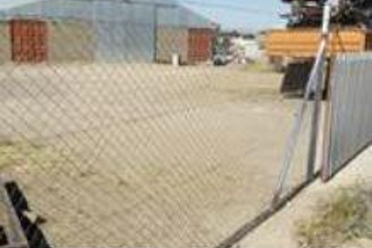 An industrial yard area featuring a large metal shed structure and perimeter chain-link fencing on an unpaved surface.