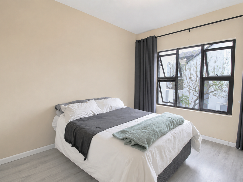 A bright bedroom featuring light-toned laminate flooring, neutral wall finishes, and large aluminium-framed windows with dark curtains.