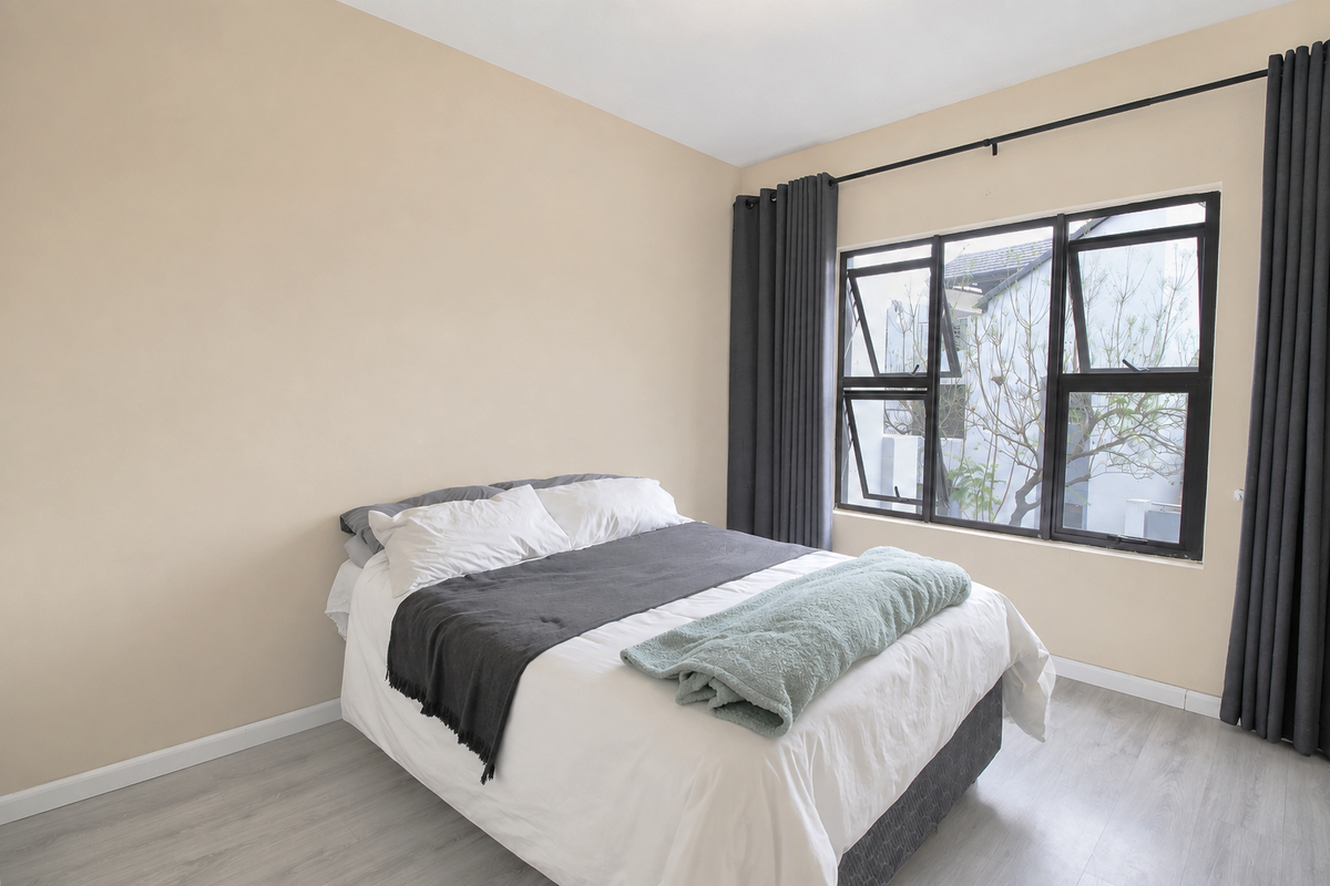 A bright bedroom featuring light-toned laminate flooring, neutral wall finishes, and large aluminium-framed windows with dark curtains.