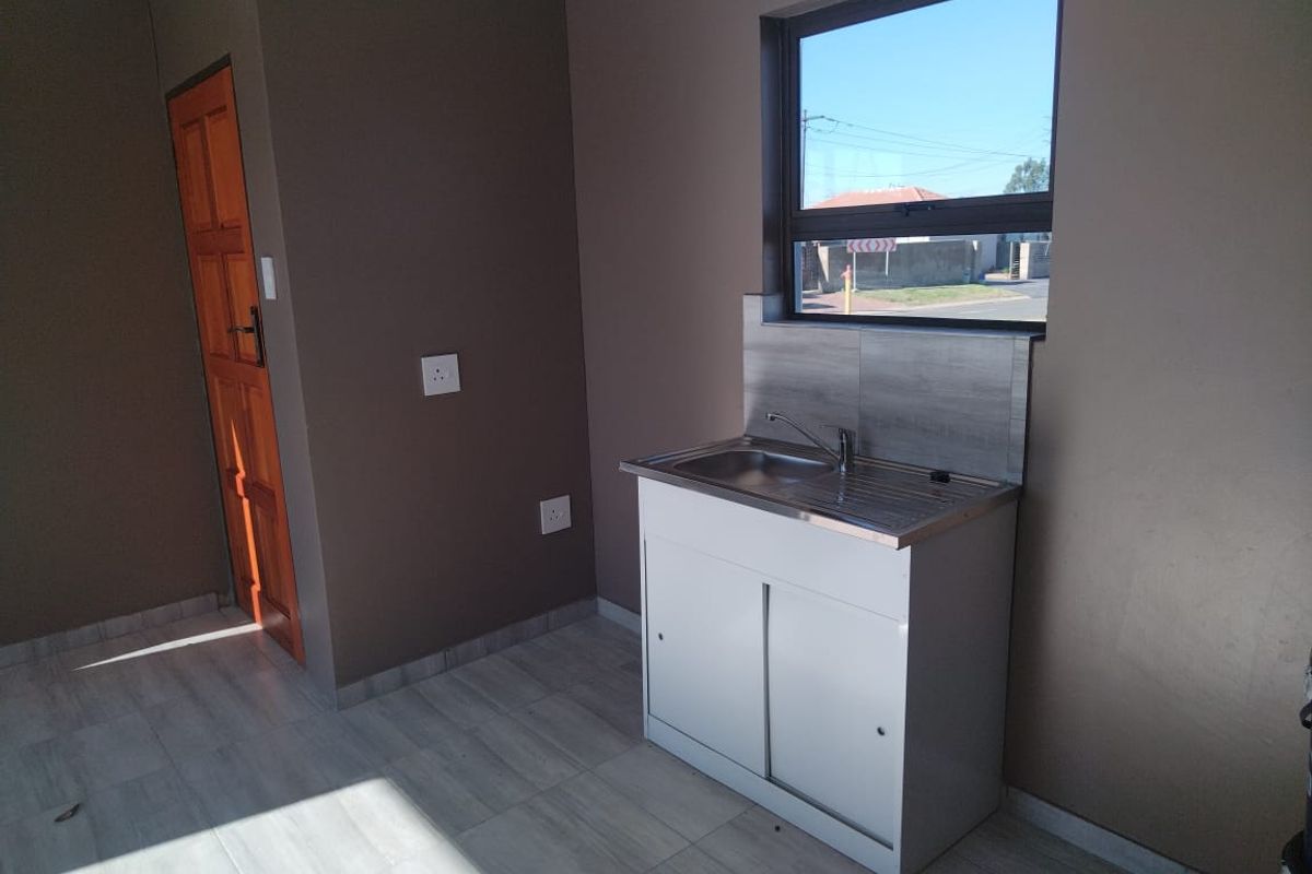 A compact, empty kitchen area featuring a stainless steel sink unit, tiled splashback, tiled flooring, and a timber door.