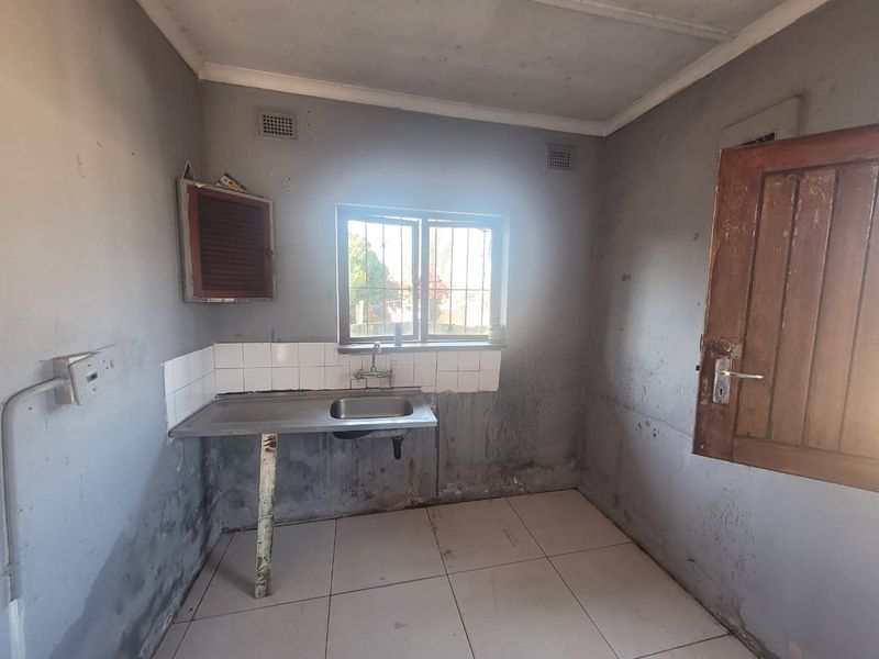 A basic, empty kitchen area featuring a stainless steel sink, tiled splashback, and tiled flooring, requiring significant cosmetic renovation and wall repairs.