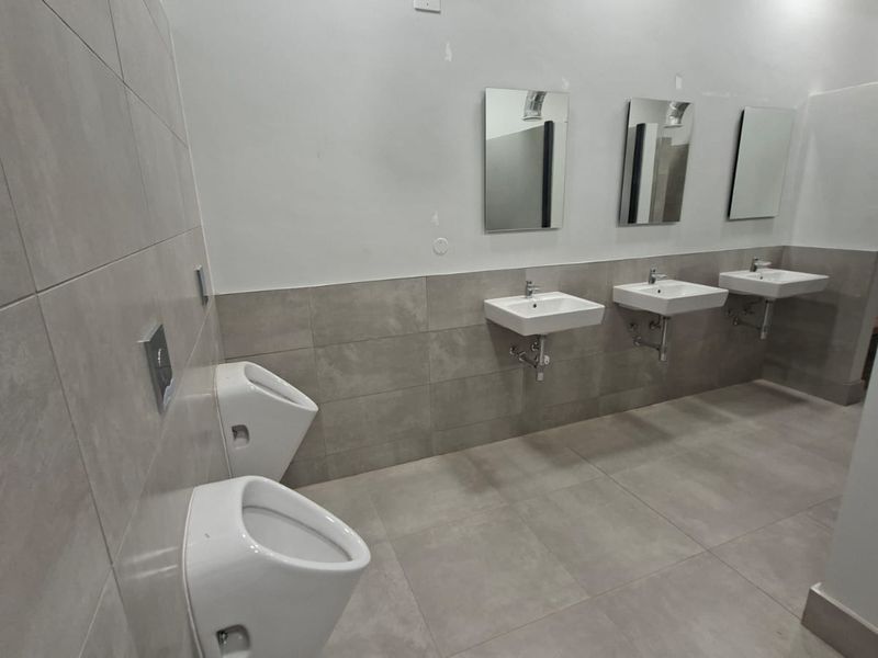 Modern industrial-style communal bathroom featuring wall-mounted urinals, multiple washbasins with chrome fixtures, and neutral large-format floor and wall tiling.